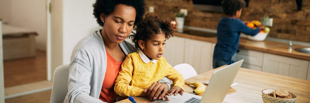 Black working mother taking notes while daughter is sitting on her lap and using laptop at home. Small boy is in the background.