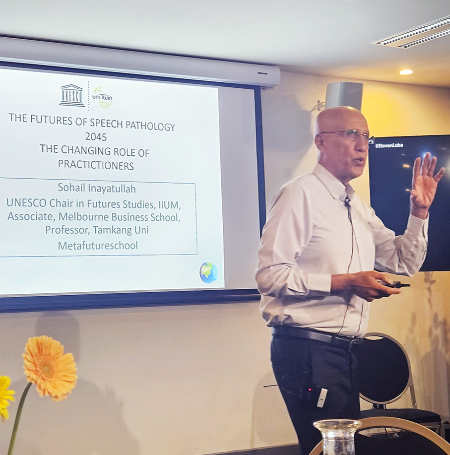 This is a photo of Professor Sohail Inayatullah at the front of the room presenting in front of a projector that is displaying his presentation. He is wearing a white shirt.  